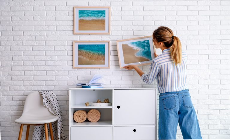 An interior decorator hangs artworks of beach scenes on a white wall in a tropical home.