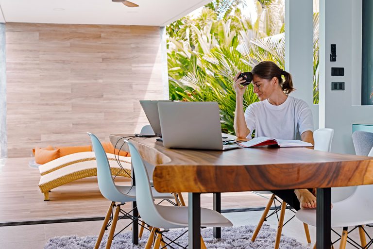 Woman sitting at table in front of open sliding doors leading onto a terrace in a tropical house.
