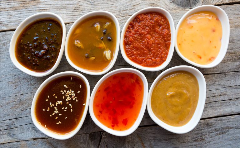 Close-up view of seven white bowls containing different Southeast Asian condiments on a wooden table.